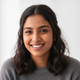 A smiling South Asian woman in her late 20s, wearing a charcoal top, with a soft terracotta glow, against a bright white back